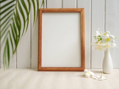 Poster ahşap çerçevesi ve güzel Frangipani tropikal çiçeği bej masa üzerindeki modern beyaz vazoda ve ahşap palmiye yaprağıyla arka planda