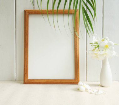 Poster ahşap çerçevesi ve güzel Frangipani tropikal çiçeği bej masa üzerindeki modern beyaz vazoda ve ahşap palmiye yaprağıyla arka planda