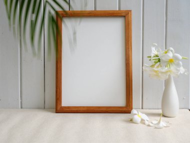 Poster ahşap çerçevesi ve güzel Frangipani tropikal çiçeği bej masa üzerindeki modern beyaz vazoda ve ahşap palmiye yaprağıyla arka planda 