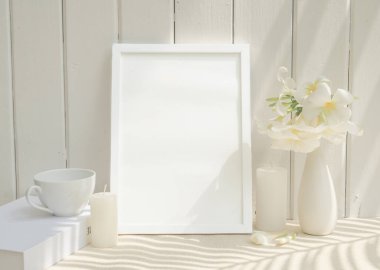 Boş model resim çerçevesi, kahve fincanı mumu ve bej masadaki seramik beyaz vazoda Plumeria çiçeği, beyaz ahşap arka plan, uzun palmiye gölgesi.
