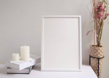 Poster çerçevesi, siyah beyaz kitaplar, kurutulmuş Lagurus ovatus çiçekleri modern cam vazo ve beyaz masa ve çimento duvar arka planında mumlar.