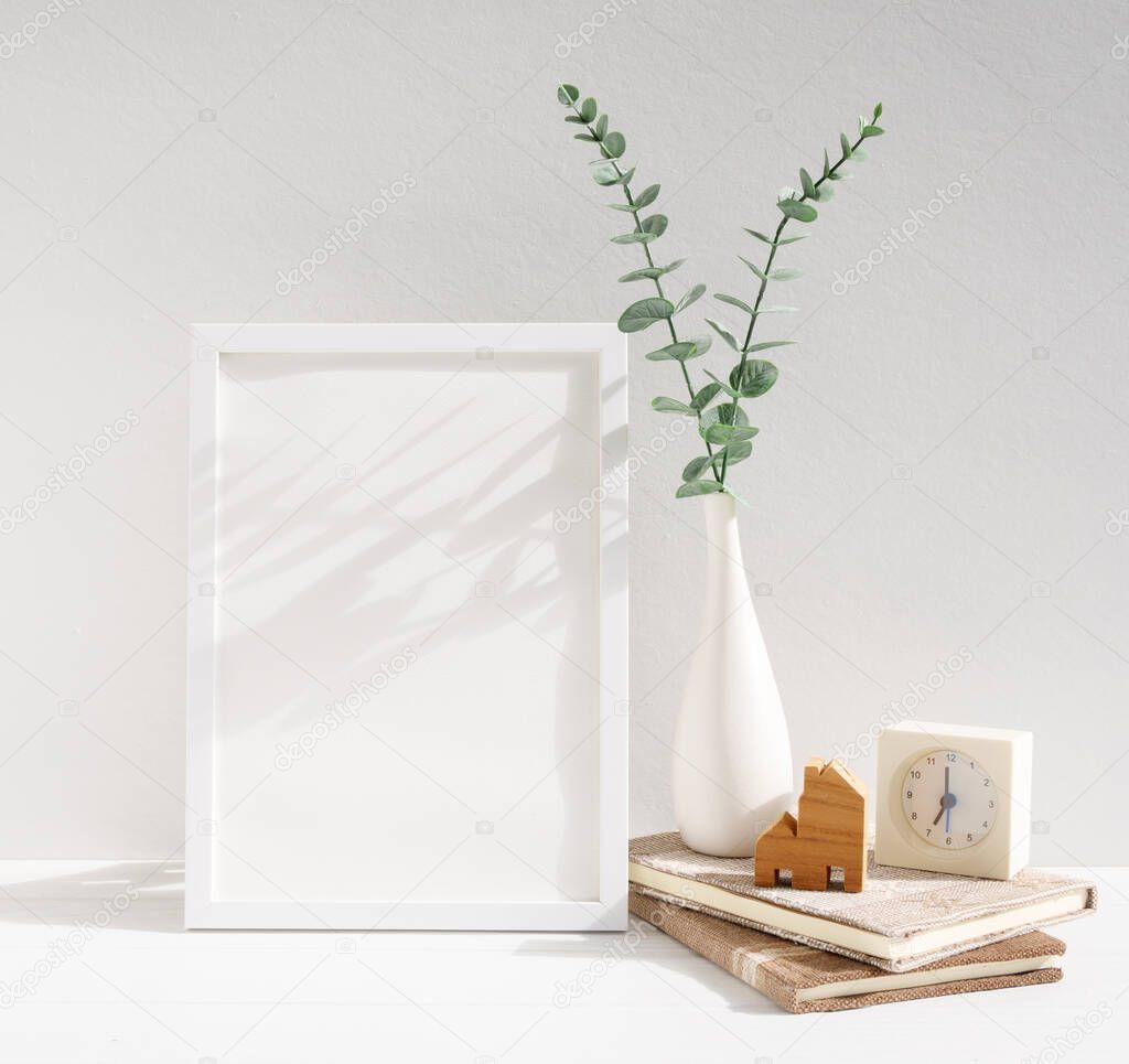 Two mock up white  poster frames decor with dried eucalyptus leaves in  modern white vase notebook,clock  and house model over white table wall background