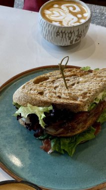 Mavi bir seramik tabakta servis edilen taze sandviç ve bir fincan latte ile birlikte sıcak bir kafede ayrıntılı latte sanatı.