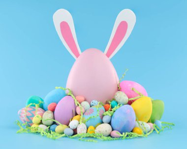 Large pink Easter egg with bunny ears surrounded by a pile of colorful Easter eggs and candy.