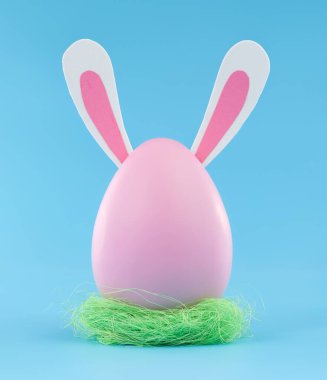 Large pink Easter egg with bunny ears in Easter grass nest. Blank space for text.