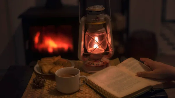 a woman reads a book and drinks tea with cookies by the light of a kerassin lamp by a burning fireplace in a cozy home atmosphere.