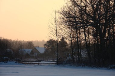 Kış sabahı. Şafak Karla kaplı çayır, ağaçların siluetleri, ev ve açık bir gökyüzü görülebilir. Resim orijinal bir kırsal çitle desteklenmiş..