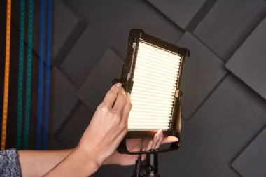 Stüdyo asistanı ayarlanabilir ışık panosundaki profesyonel video ışığını tamir etti. Fotoğraf stüdyosunda üç ayaklı lamba. Kabartmalı gri duvar arkaplanındaki fotoğraf ve video lambası