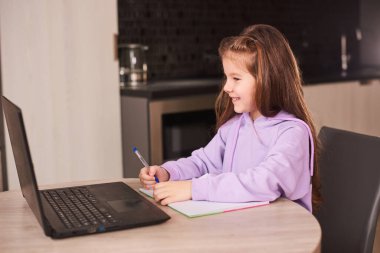 Okullu kız mutfakta ödev yapıyor, bilgisayar uygulaması kullanarak video dersi izliyor, evde sanal derslerle ilgileniyor.