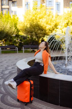 Bavulda oturan kadın şehir parkında tatilin tadını çıkarıyor.