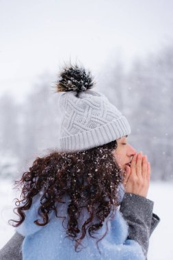 Kadın donmuş parmaklarını ısıtıyor ve kar arka planında avucuna nefes alıyor.