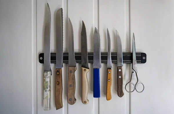 Un ensemble de couteaux de chefs avec différentes poignées et différentes longueurs sont suspendus sur un tampon magnétique sur un mur blanc plus des ciseaux — Photo
