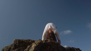 Genç beyaz kadın esmer kayalık sahilde mavi gökyüzüne karşı yoga yapıyor.