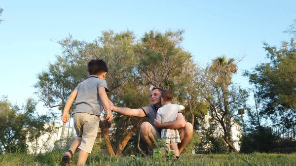 Un padre joven en el parque llamando a sus hijos. Disparo de ángulo bajo. Chicos corriendo hacia papá —  Fotos de Stock