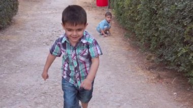 Parkta oynayan iki çocuk. Yürüyen çocuk çalılara bakıyor ve anaokulu çocuğu kameraya doğru koşuyor.