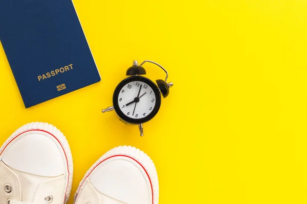 Pasaport, çalar saat ve sarı arka planda sakız ayakkabıları. Seyahat zamanı. Seyahat aksesuarları. Tatil ve tatil konsepti. Üst görünüm, kopyalama alanı olan düz uzanma.