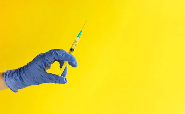 Sarı arka planda plastik şırınga tutan mavi eldivenli doktorun yakın çekimi. Tıbbi enjeksiyon. Covid-19 aşısı, sağlık hizmeti, tedavi konsepti.