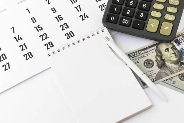 Kalem, takvim, hesap makinesi ve 100 dolarlık banknotlarla dolu boş bir defter. Muhasebe veya muhasebe, finans borcu, vergi ödemesi, iş konsepti. 