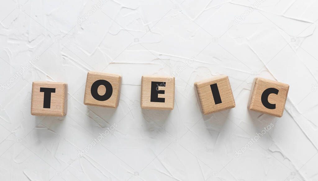 Text TOEIC on wooden cubes on white textured putty background. Abbreviation of 'Test of English for International Communication'. Test of English language. Square wood blocks. Top view, flat lay.