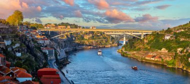 Porto, Portekiz. Douro nehrinde köprü ve sahil şeridindeki eski kasabaların evleri görünüyor. Gün, mavi gökyüzü ve bulutlu manzara manzarası. Nehirde tekneler