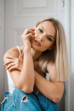 Güzel bir kadın beyaz kapının yanında otururken poz veriyor.