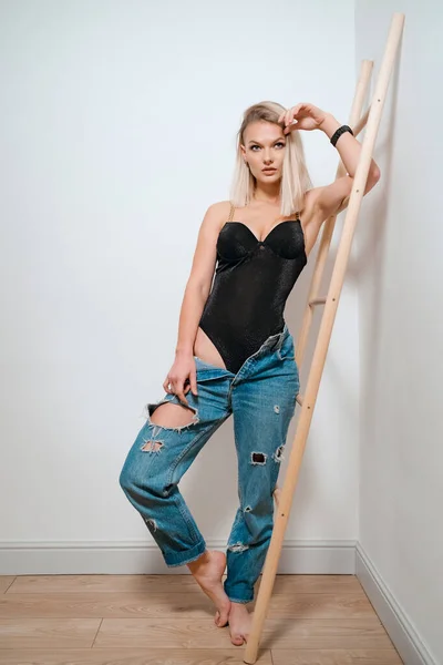 Блондинка в черном теле и джинсах позирует на фоне стены — стоковое фото