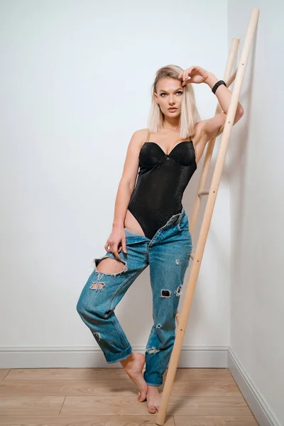 Mujer rubia en cuerpo negro y jeans posando sobre fondo de pared —  Fotos de Stock