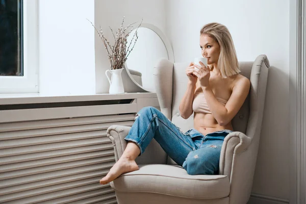 Vrouw in beige beha en blauwe jeans die thuis koffie drinkt — Stockfoto