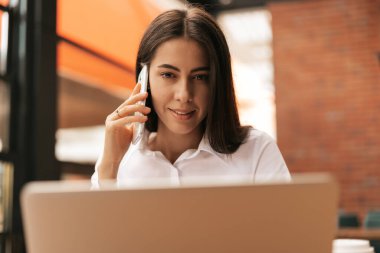 Kurumsal İletişim. Kafkasyalı iş kadını cep telefonuyla konuşuyor. Modern ofiste dizüstü bilgisayarla çalışıyor..