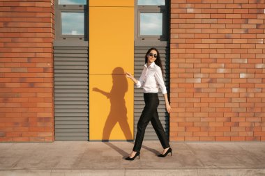 Yüksek tepelerdeki genç bir kadın güneş gözlükleriyle güneşli bir günde tuğla bina duvarında yürürken ayakkabılarını ve iş kıyafetlerini giyiyor.