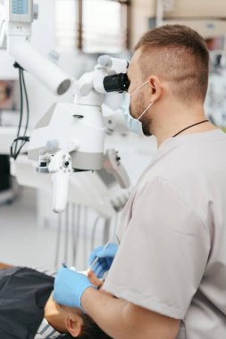 Erkek dişçi maske takıyor. Mikroskoba bakıyor.