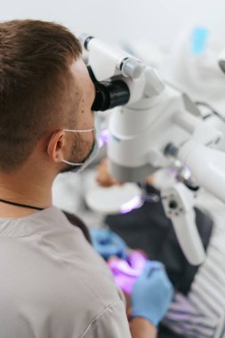 Erkek dişçi maske takıyor. Mikroskoba bakıyor.