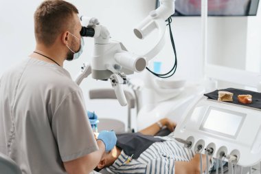 Erkek dişçi maske takıyor. Mikroskoba bakıyor.