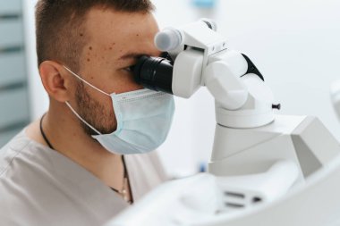 Erkek dişçi maske takıyor. Mikroskoba bakıyor.