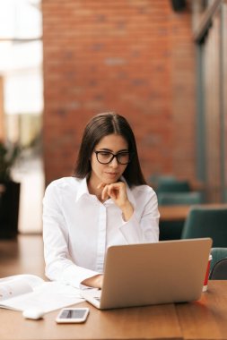 Gözlüklü güzel bir kadın oturmuş çalışırken dizüstü bilgisayarla konuşuyor.