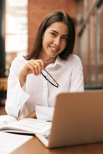 Hermosa mujer en gafas hablando con el ordenador portátil con sonrisa mientras está sentado y trabajando — Foto de Stock