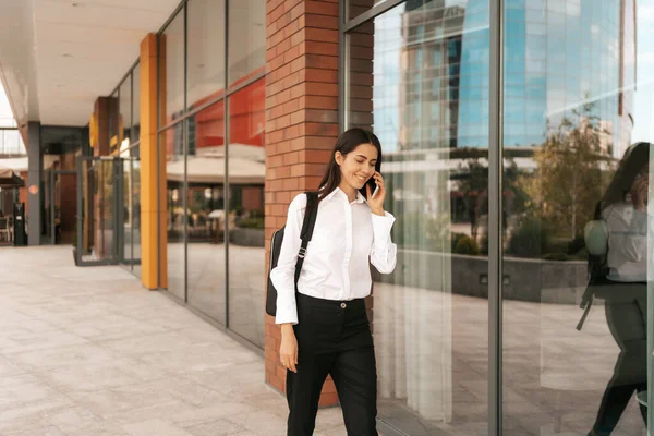 Woman with business backpak walking while speaking on the phone near a a business building