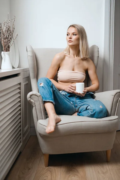 Γυναίκα σε μπεζ σουτιέν και μπλε τζιν πίνοντας καφέ στο σπίτι — Φωτογραφία Αρχείου
