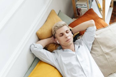 Genç adam kanepede dinlenirken kulaklıkla müzik dinliyor.