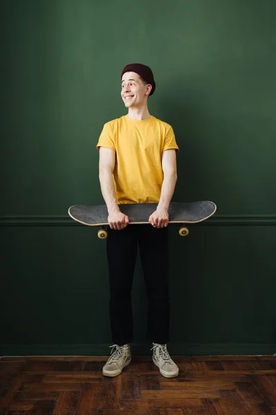 Hombre caucásico en camiseta amarilla posando con patín sobre pared de fondo verde oscuro —  Fotos de Stock