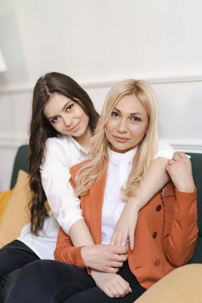 Bela mãe e sua filha adulta estão abraçando e sorrindo enquanto estão sentados no sofá em casa — Fotografia de Stock
