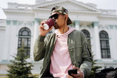 Parkta güneşli bir günde kahve içerken akıllı telefon kullanan yakışıklı adam.