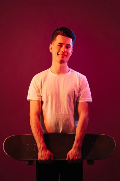 Caucasian man in white t shirt posing with skate over dark crimson background wall — Stock Photo, Image