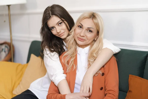 Die schöne Mutter und ihre erwachsene Tochter umarmen sich und lächeln, während sie zu Hause auf der Couch sitzen — Stockfoto