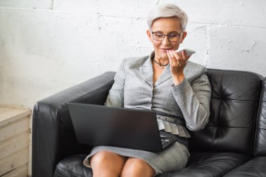 Takım elbiseli güzel, şık, yetişkin bir kadın. Kanepede oturuyor. Laptop ve telefonla.