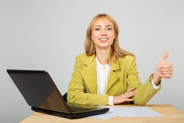 漂亮的年轻女性 身穿夹克 手握手提电脑 站在桌边 摆出一副手势 — 图库照片