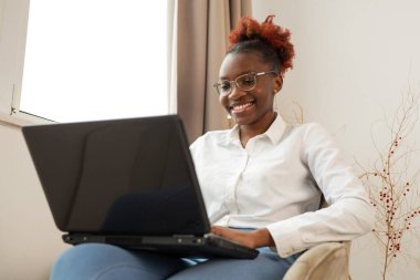 Güzel, genç bir Afrikalı kadın, elinde dizüstü bilgisayarla pencerenin yanında oturuyor.