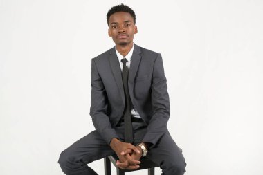 Beyaz arka planda, koltuğunda oturan takım elbiseli yakışıklı Afrikalı genç adam.