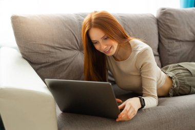 Kızıl saçlı güzel bir bayan evde dizüstü bilgisayarla kanepede yatıyor.