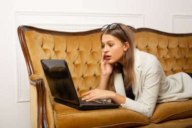 Güzel, genç bir bayan dizüstü bilgisayarla kanepede yatıyor. Şaşırmış bir yüzü var.